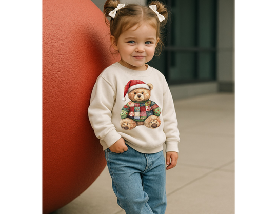 Toddler in Christmas Bear Toddler Crew sweatshirt, featuring a patchwork teddy bear in a Santa hat, stands against a red sphere, smiling sweetly with hair in buns.