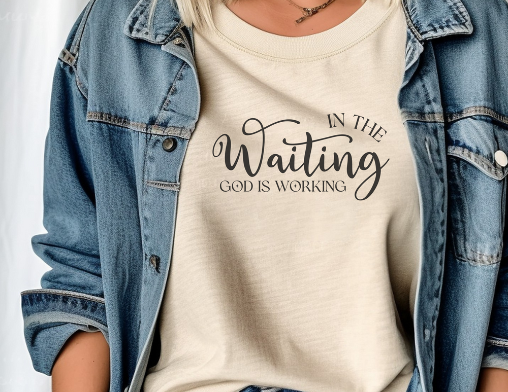 A woman in a denim jacket, showcasing the God is Working Tee from Worlds Worst Tees. A comfortable, ring-spun cotton t-shirt with a relaxed fit and durable stitching, perfect for daily wear.