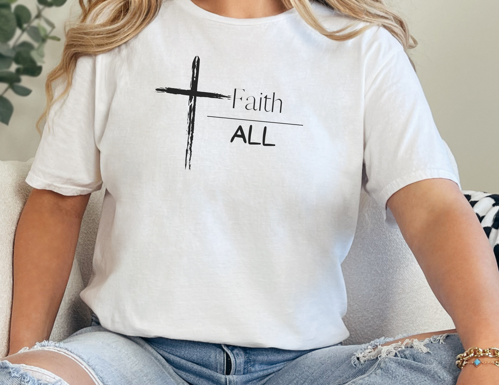 A woman in a white shirt with a cross, sitting on a chair. Faith Over All Tee: 100% ring-spun cotton, medium weight, relaxed fit, durable double-needle stitching, tubular shape.