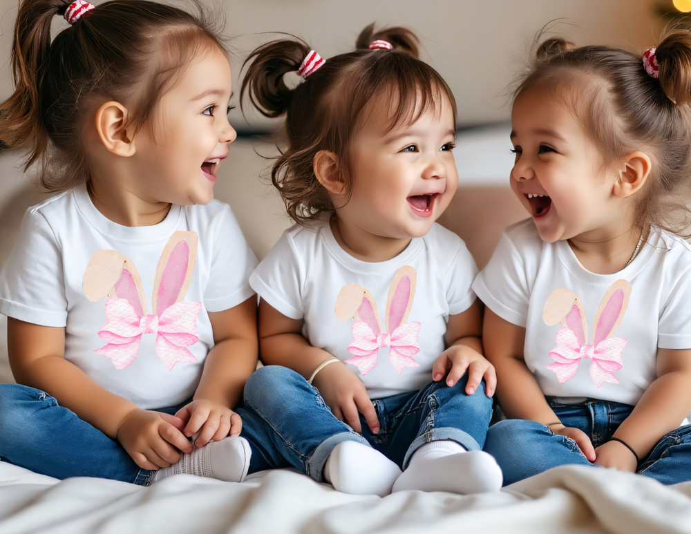 Three toddlers in Bunny Bow Baby Tees with pastel pink bunny ears, blue jeans, and ponytails, sitting joyfully together on a couch.