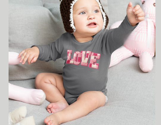 Infant wearing a LOVE onesie with a knit cap, sitting on a couch surrounded by toys, reaching upward.