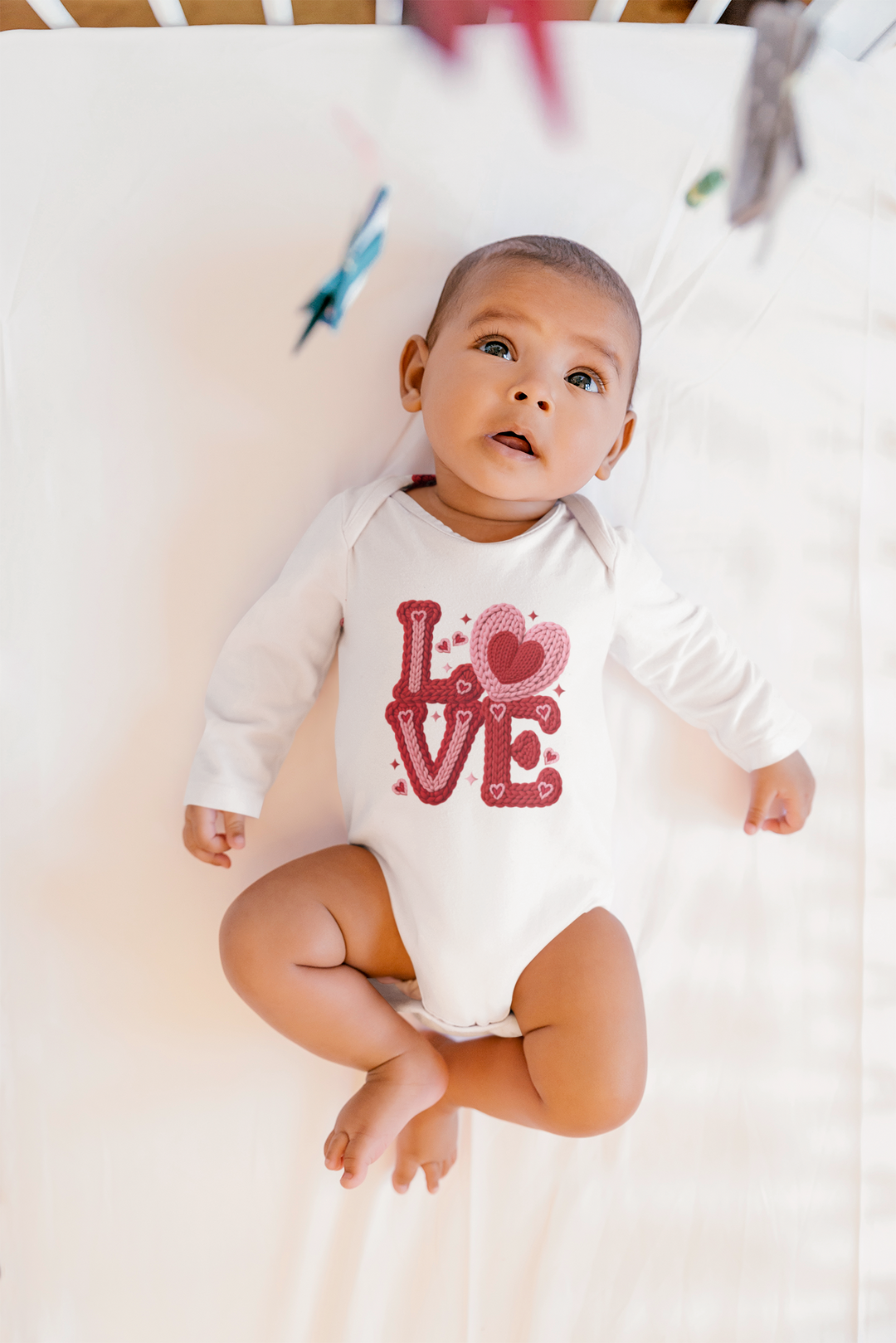 Infant in crib wearing the Love Heart Onesie, featuring LOVE knitted in red and pink. Soft, durable cotton fabric with convenient snap closure for easy changing.