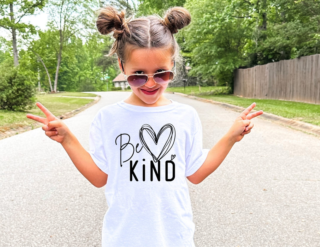 Young girl wearing a Be Kind Kids Tee smiles with peace signs in a tree-lined street, showcasing the tee's comfort and positive message.
