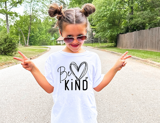 Young girl wearing a Be Kind Kids Tee smiles with peace signs in a tree-lined street, showcasing the tee's comfort and positive message.