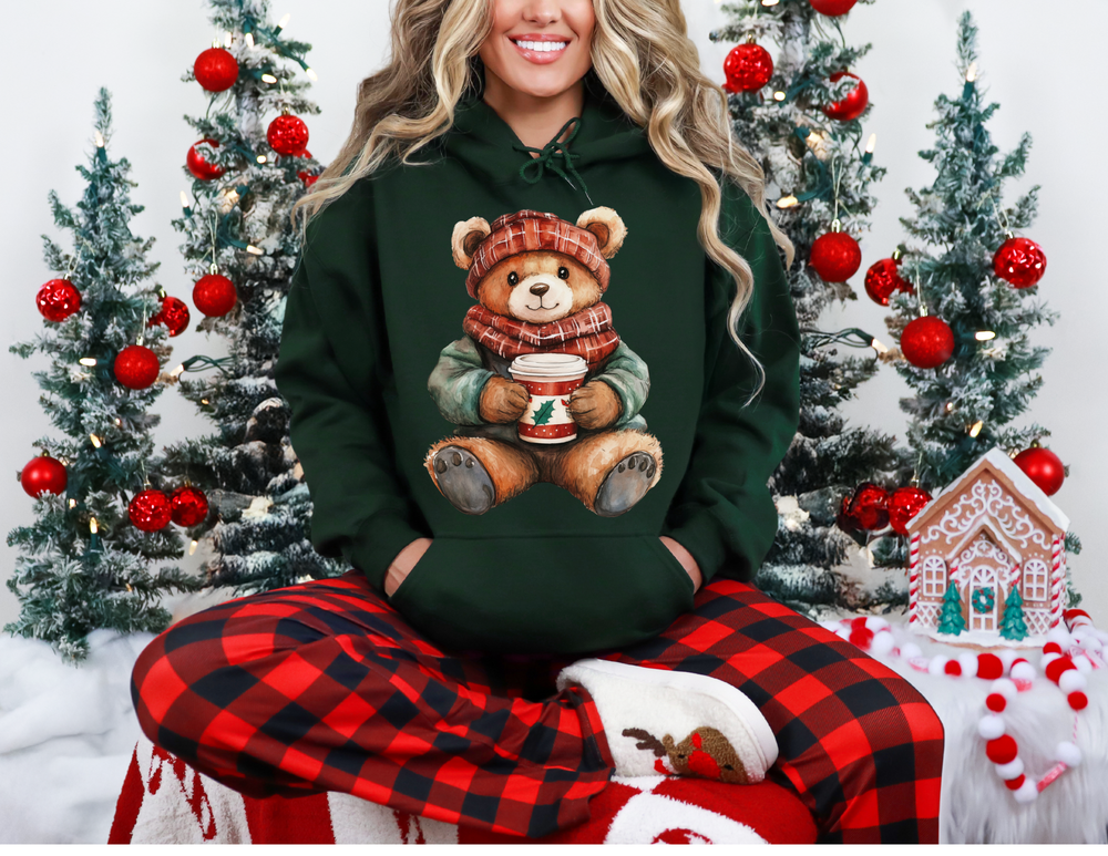 Person wearing Old Money Bear Hoodie with teddy bear design, red-and-black plaid pajama pants, and fuzzy slippers, surrounded by festive decor and lit evergreen trees.