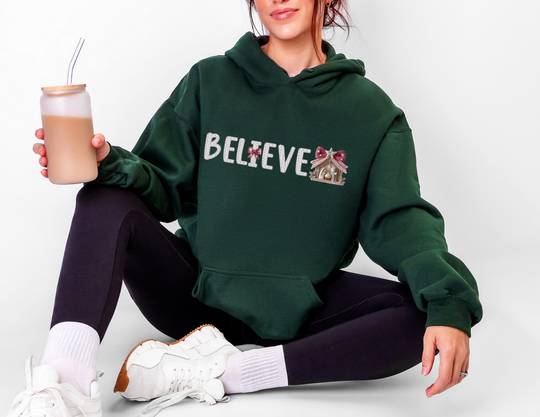 Person wearing a Believe hoodie with nativity-themed lettering, holding iced coffee, seated cross-legged on the floor, embodying cozy, stylish comfort.