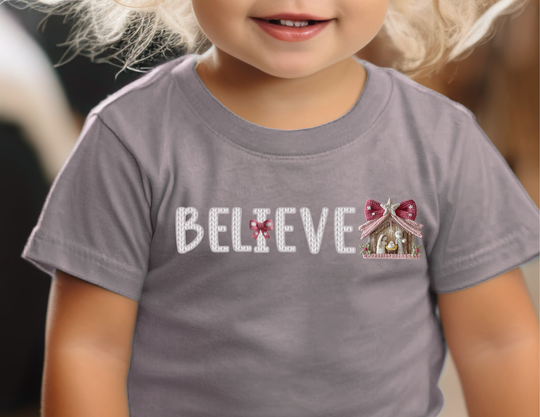 Young child wearing the Believe Baby Tee with knitted-style letters, pink ribbon, and nativity scene. Soft, durable, perfect for baby's first steps.