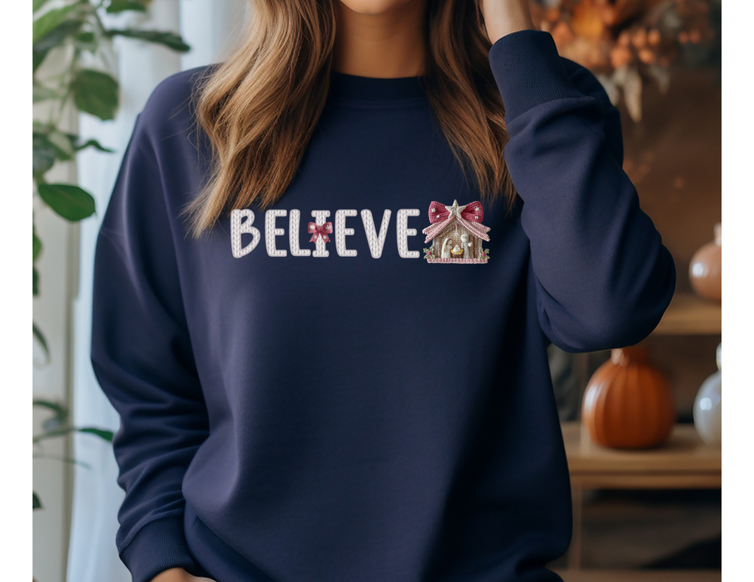 Navy sweatshirt with BELIEVE in knit-stitch style, featuring a red bow and nativity stable, embodying cozy autumn-to-holiday vibes.