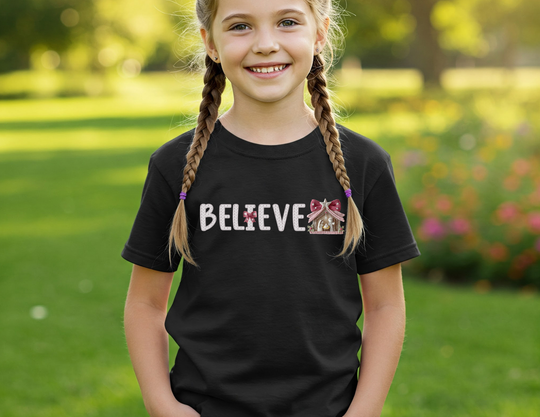 Young girl in Believe graphic tee with braided hair, smiling outdoors on a sunny day. The T-shirt features playful details with bows and a gingerbread-house icon.
