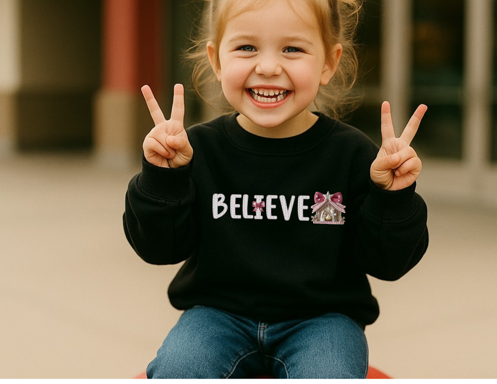 Child wearing BELIEVE toddler sweatshirt, smiling with peace signs, showcasing a playful nativity scene design on the letter E. Cozy, durable for warmth and comfort.