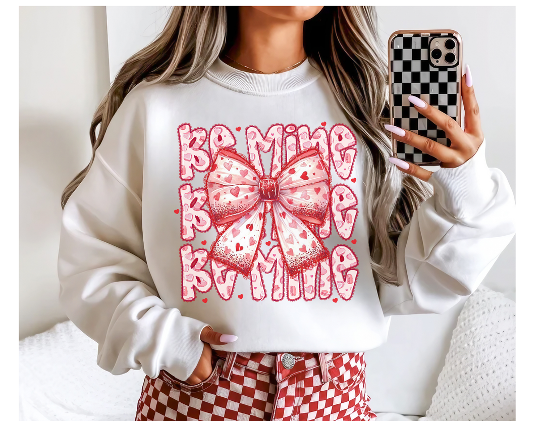 Person wearing Be Mine Crew sweatshirt with pink-and-red bow design, taking a mirror selfie with checkerboard phone case. Red-and-white pants visible in the background.
