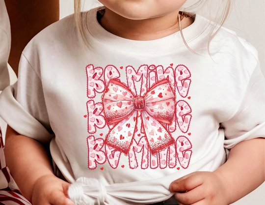 Child wearing Be Mine Kids Tee with pink-and-red bow and MAMA pattern, highlighting the tee's playful graphic design and everyday comfort from Faith Family Designs.