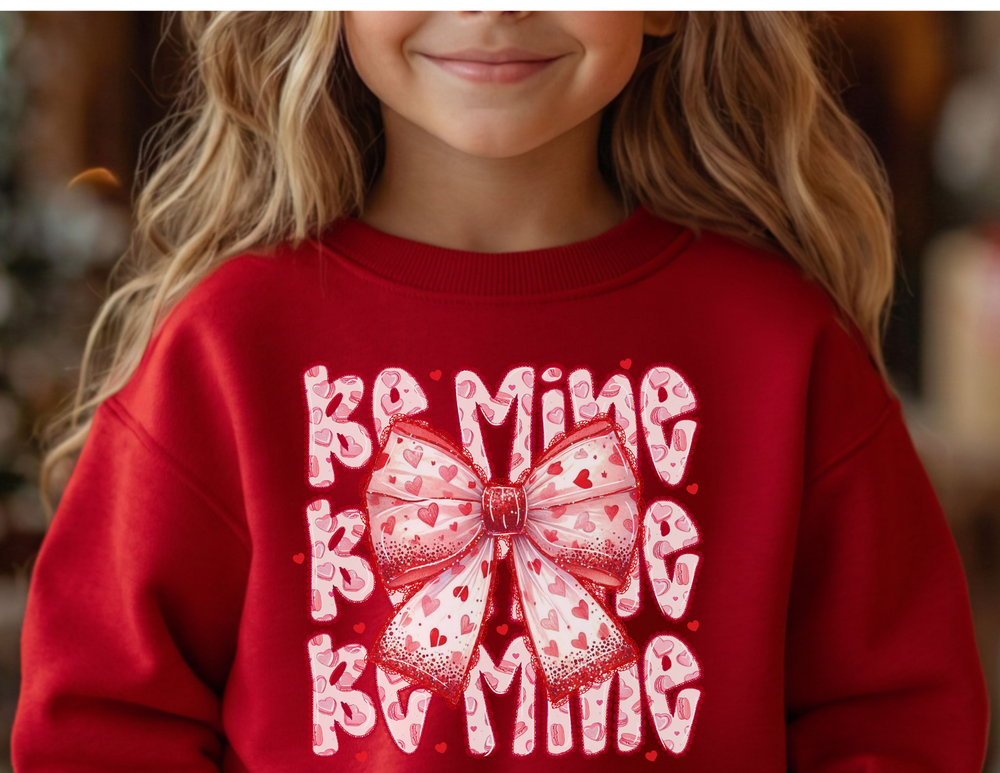 Young girl wearing a Be Mine Toddler Crew sweatshirt with a large pink bow and heart-spotted letters, embodying a playful Valentine's Day theme.