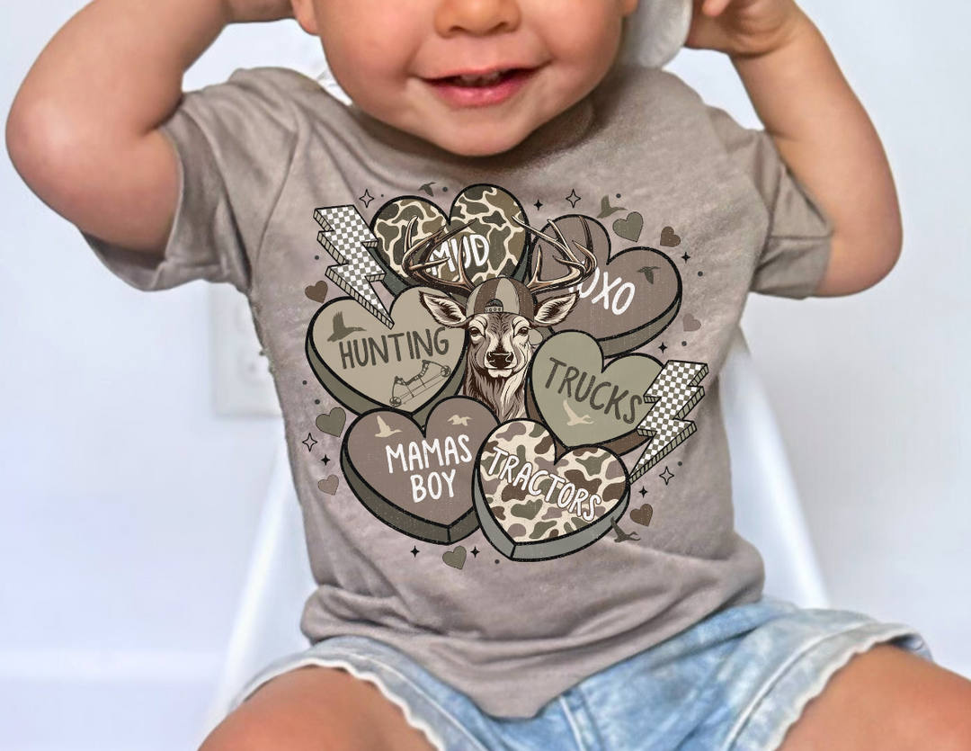 Toddler wearing a beige Boy Valentine Baby Tee with a stylized deer head and camouflage hearts, seated in a white chair, showcasing playful graphics.