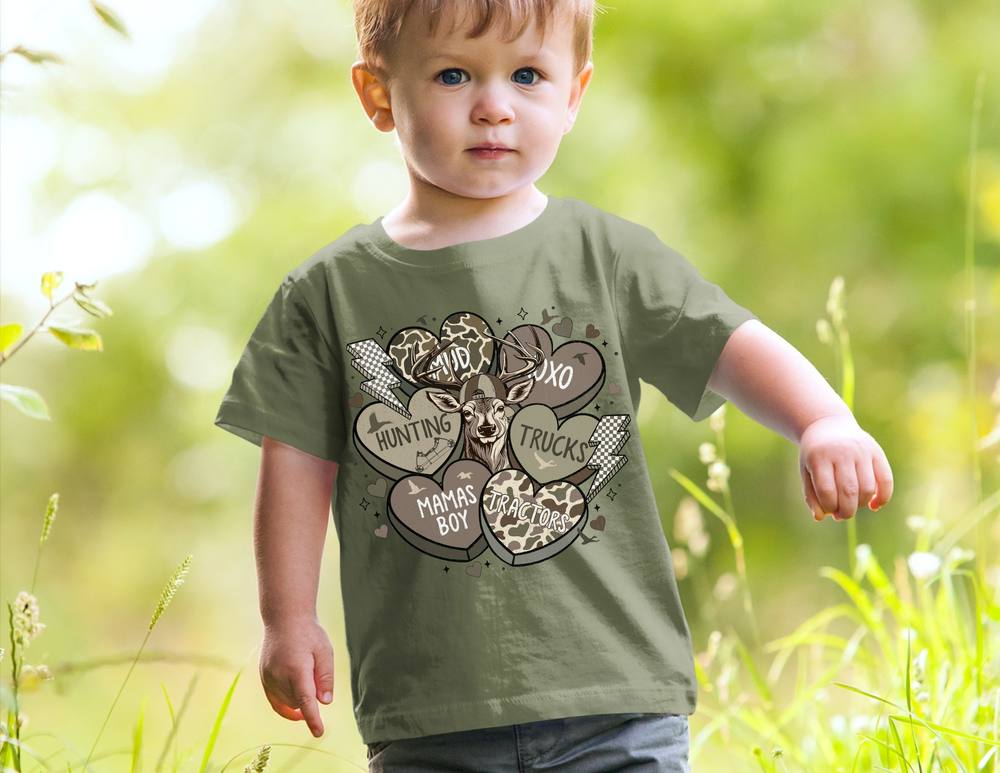 Toddler wearing Boy Valentine Toddler Tee with a deer head print and playful designs, standing outdoors in a grassy, sunlit area.