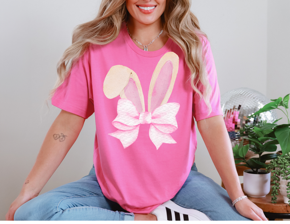 Person wearing Bunny Bow Tee with bunny ears graphic, light-washed jeans, and white sneakers. Nearby, a disco ball and plants on a side table.