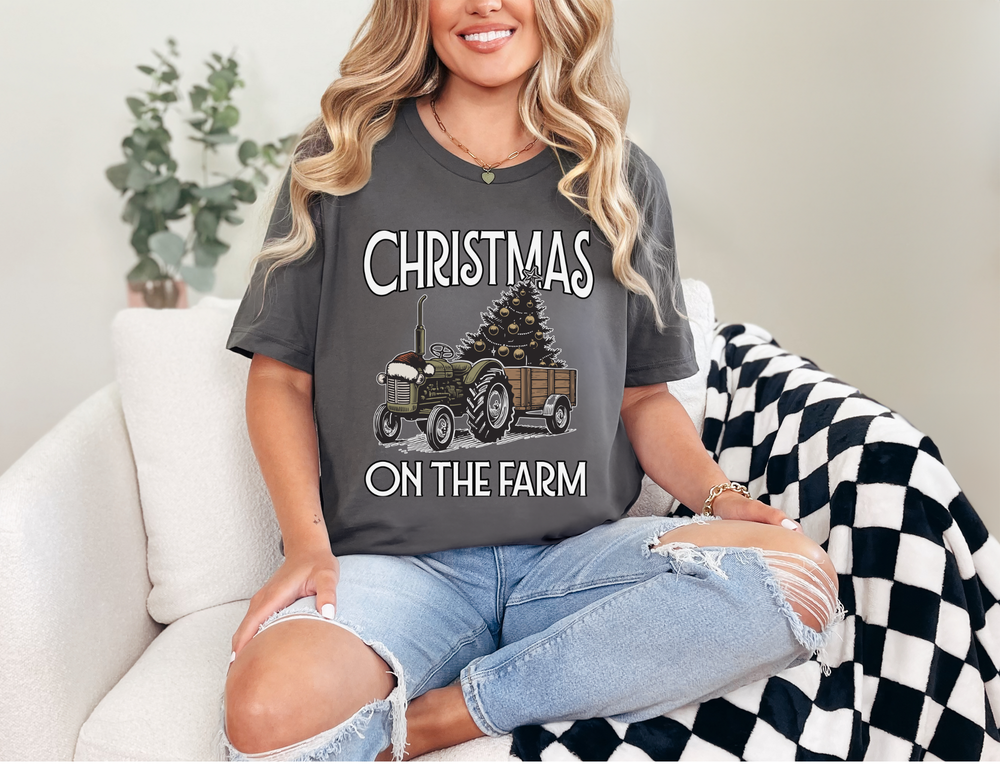Woman in Christmas on the Farm tee with tractor and tree graphic, sitting on sofa with checkered blanket; casual, festive style.