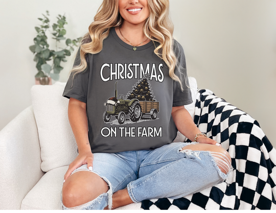 Woman in Christmas on the Farm tee with tractor and tree graphic, sitting on sofa with checkered blanket; casual, festive style.