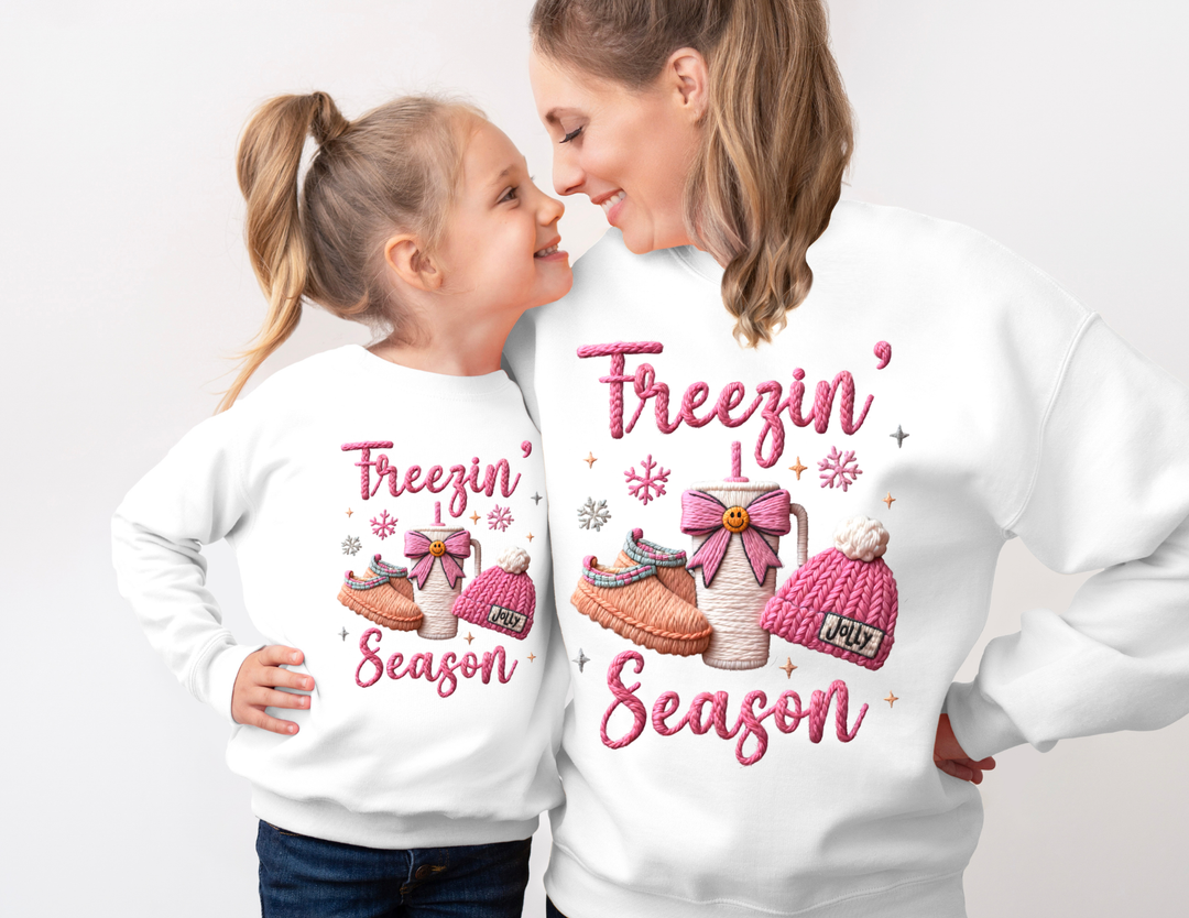 Mother and daughter wearing matching Freezin’ Season white sweatshirts with winter motifs, showcasing cozy toddler apparel from Faith Family Designs.