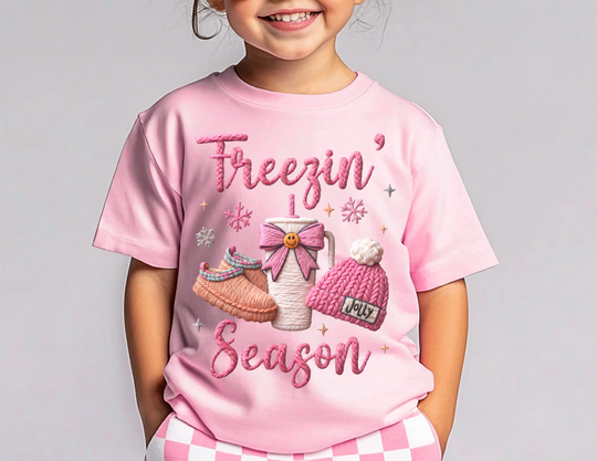 Child wearing Freezin Season Kids Tee with winter-themed design, featuring a steaming mug, knitted slippers, and beanie. Hands in pink checkerboard pants pockets.