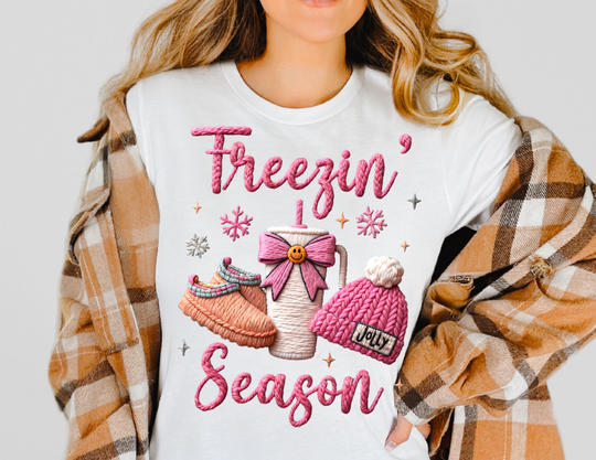 Freezin' Season Tee: White T-shirt with colorful winter-themed embroidery, featuring Freezin’ Season in pink and cozy winter items, worn under a plaid jacket.