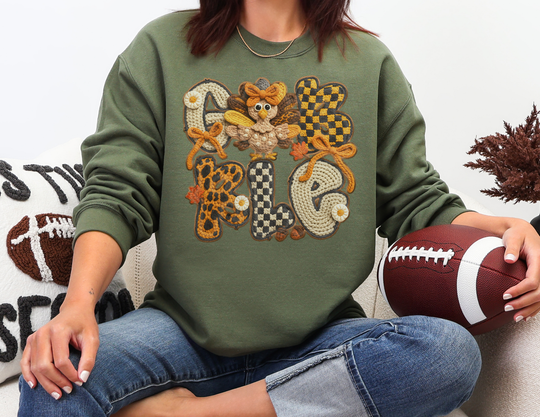 Person wearing a Gobble Crew olive-green sweatshirt with autumnal patterns, holding a football, showcasing playful design ideal for cozy, festive occasions.