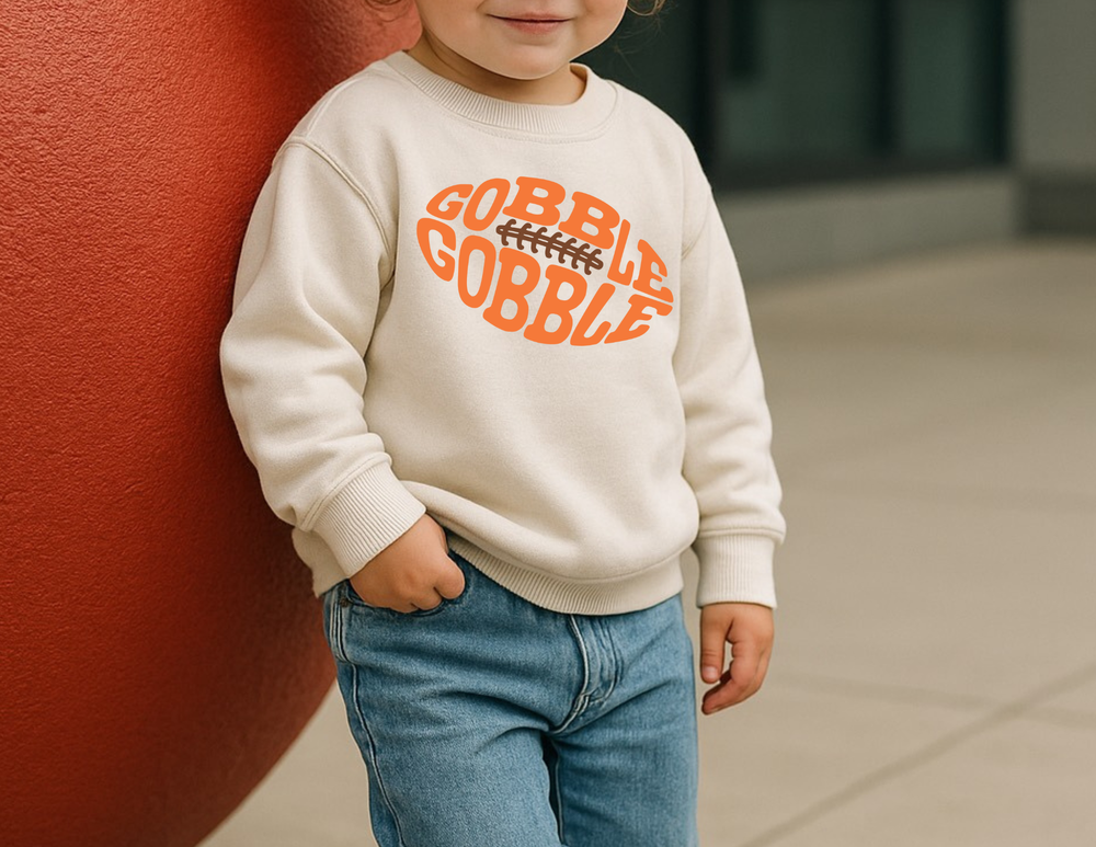 Child in light-wash jeans wears a cream GOBBLE GOBBLE football-themed sweatshirt, highlighting comfortable, durable design for toddlers from Faith Family Designs.