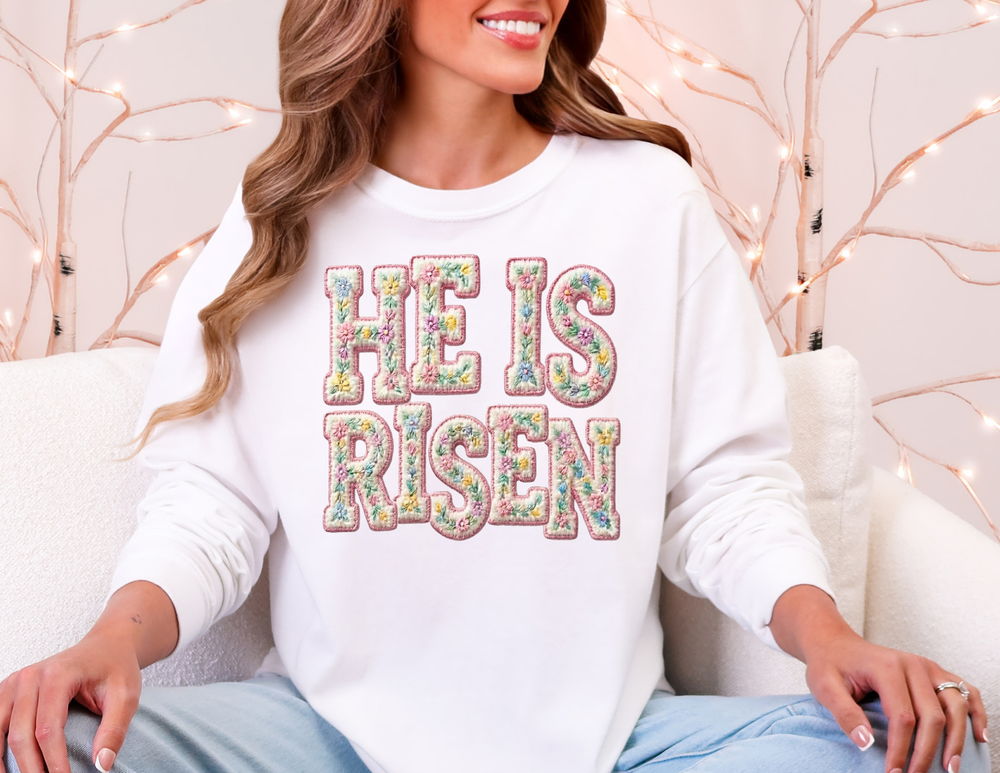 Woman wearing He is Risen long sleeve tee with floral embroidery, seated on a light sofa, surrounded by warm fairy lights, embodying a cozy, festive atmosphere.