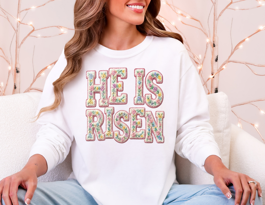 Woman wearing He is Risen long sleeve tee with floral embroidery, seated on a light sofa, surrounded by warm fairy lights, embodying a cozy, festive atmosphere.