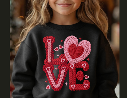 Young girl wearing the Love Heart Toddler Crew sweatshirt, featuring a knitted-style LOVE graphic with overlapping heart-shaped O, surrounded by tiny hearts and sparkles.