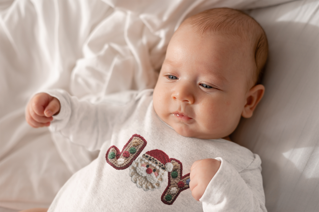 Infant in a Joy onesie with Santa motif, lying on a white blanket, highlighting soft cotton fabric and durable snaps for easy changing.