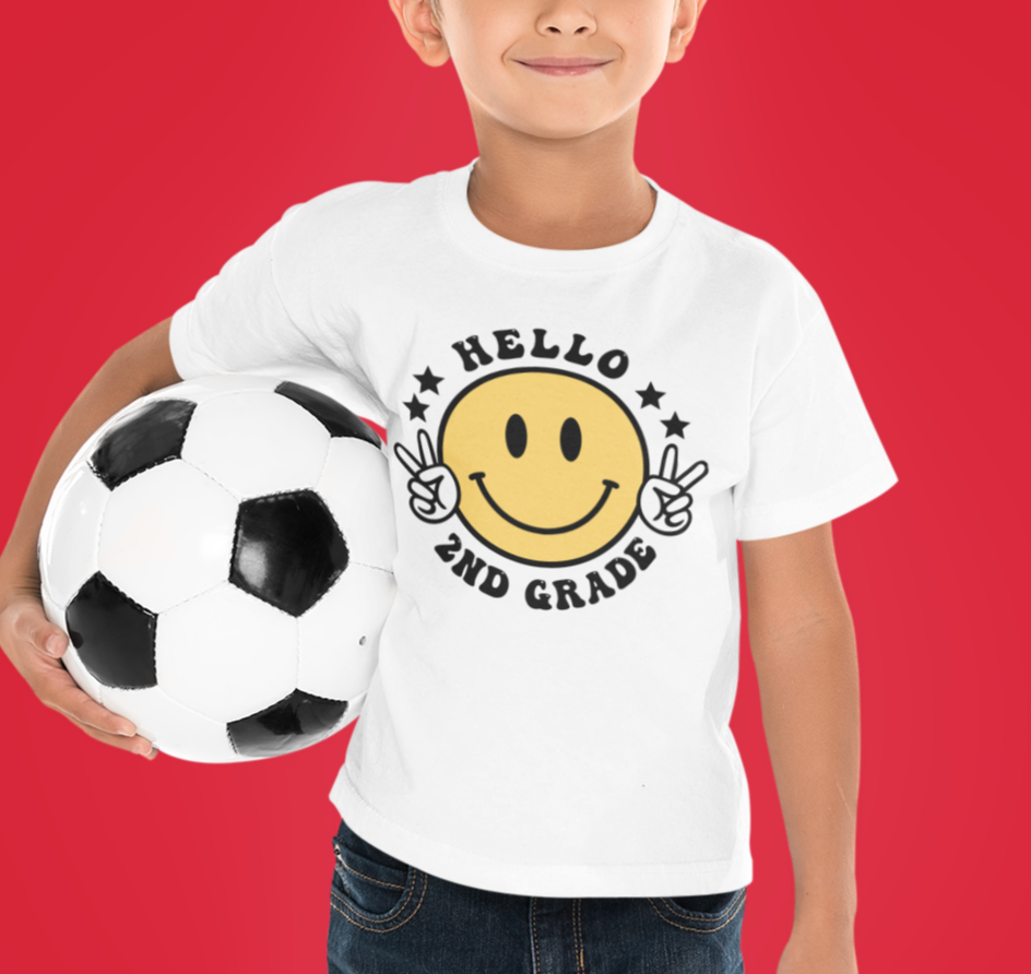 A boy holding a football ball, wearing a Hello 2nd Grade Kids Tee from Worlds Worst Tees. Made of 100% cotton, light fabric, classic fit, tear-away label, and durable twill tape shoulders.