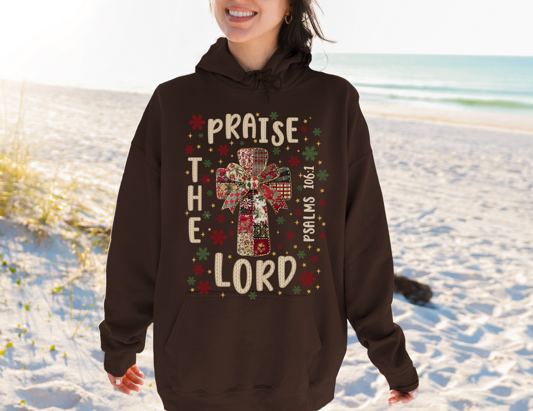 Person on beach wearing Praise the Lord Hoodie with quilt-style cross, stars, snowflakes, and Psalm 106:1, showcasing Faith Family Designs' unique graphic apparel.