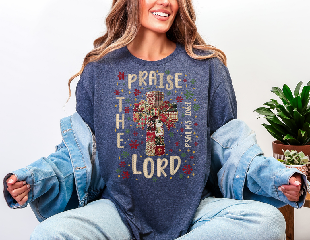 Woman in heather-navy Praise the Lord Tee with patchwork cross, festive bow design, paired with light jeans and a denim jacket draped off her shoulders.
