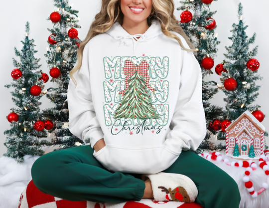 Person in a Merry Christmas hoodie with a stylized tree, seated amidst festive decor, showcasing cozy holiday attire with green joggers and reindeer slipper socks.