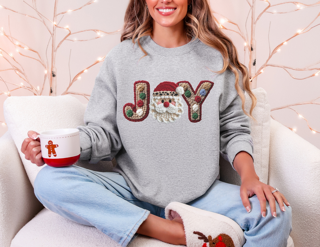 Woman in JOY crewneck sweatshirt with Santa motif, holding a festive mug, embodies cozy holiday comfort on a light sofa.