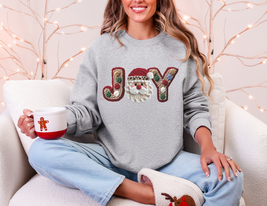 Woman in JOY crewneck sweatshirt with Santa motif, holding a festive mug, embodies cozy holiday comfort on a light sofa.
