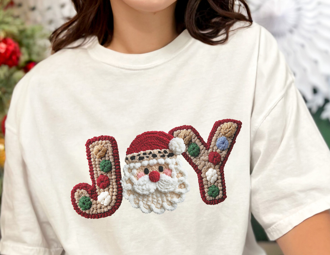 Person wearing the Joy Tee featuring a textured holiday design with JOY in gingerbread style, Santa-face motif, and colorful pom-pom accents on an off-white T-shirt.