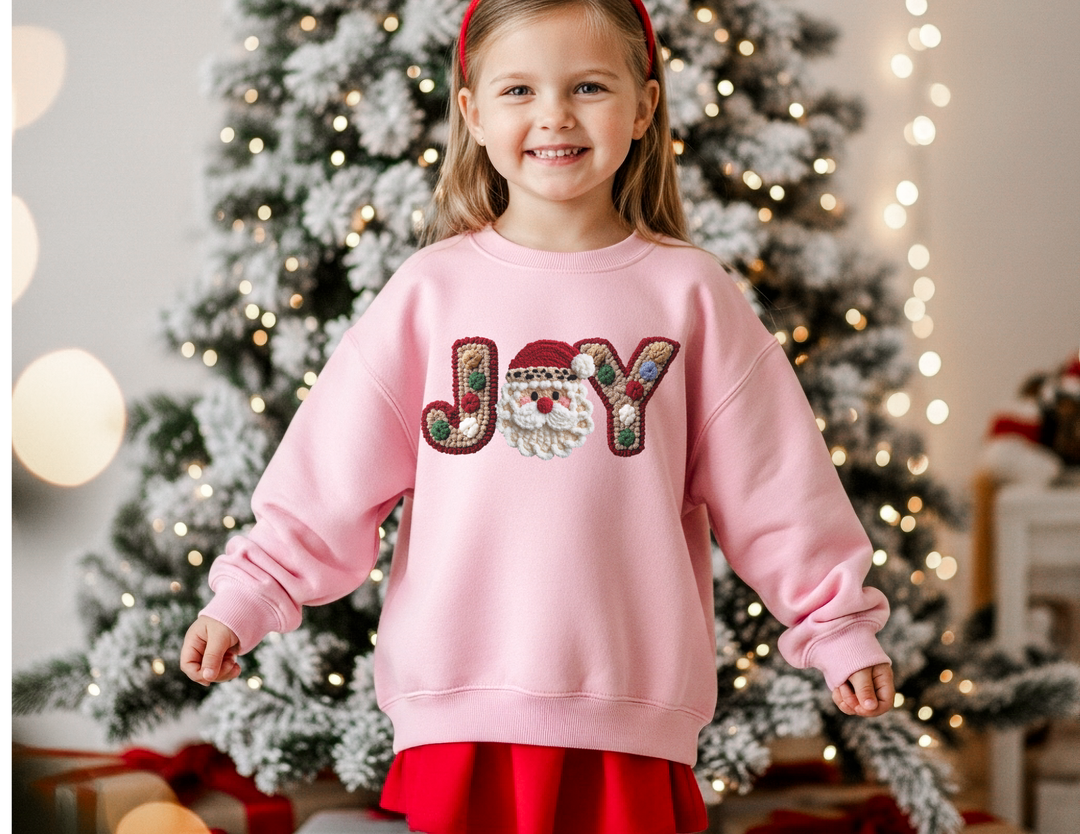 Young girl in a JOY sweatshirt with Santa motif, standing by a lit Christmas tree with presents, capturing festive warmth and comfort.
