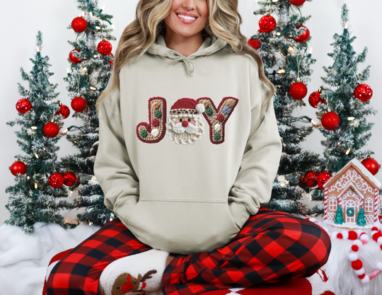 Person wearing a beige JOY hoodie with a Santa motif, buffalo-plaid pajama pants, and reindeer slippers, surrounded by festive Christmas decor.