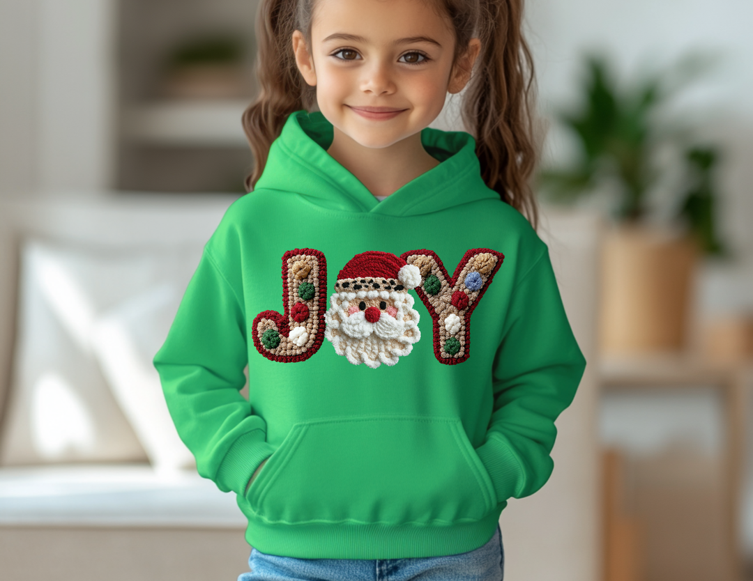 Young girl wearing the Joy Youth Hoodie with JOY and Santa motif, featuring pom-pom accents, in a cozy, softly lit living room.