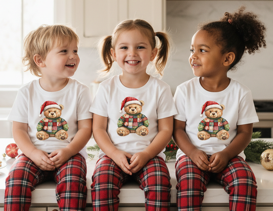 Children wearing Christmas Bear Kids Tee with teddy bear design and plaid pajama pants, sitting on a kitchen bench surrounded by holiday decor.