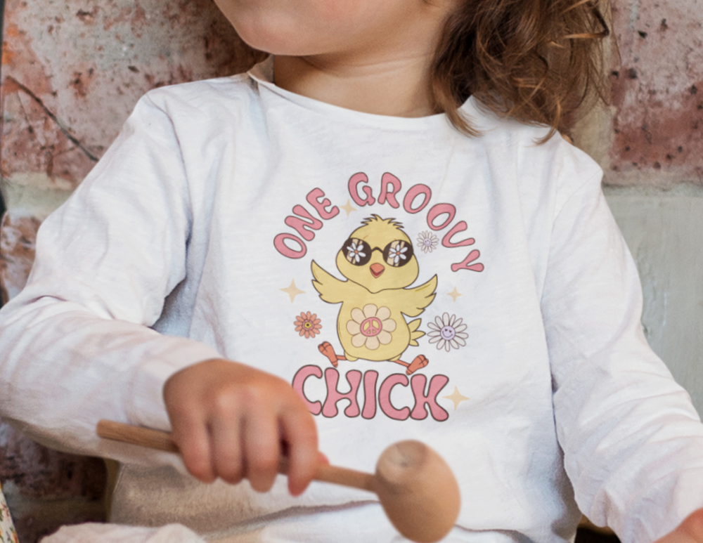 A toddler holds a wooden spoon, embodying the playful spirit of the One Groovy Chick Toddler Tee by Worlds Worst Tees. Classic fit, durable ringspun cotton tee for sizes 2T-6T.