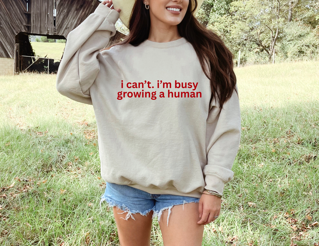 A woman in a field wears the Growing a Human Crew sweatshirt, showcasing its cozy, durable design with red text, paired with denim shorts and a straw hat.
