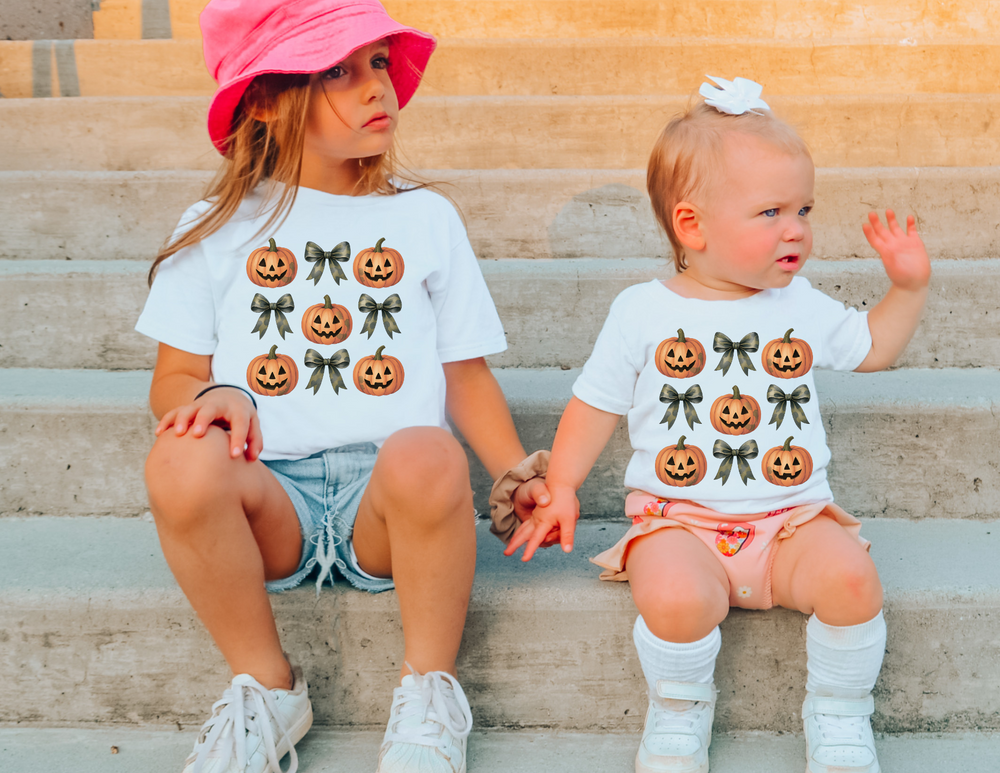 Children wearing Pumpkins and Bows Baby Tee sit on steps, featuring jack-o’-lantern designs. The older child wears a pink hat; the toddler waves.