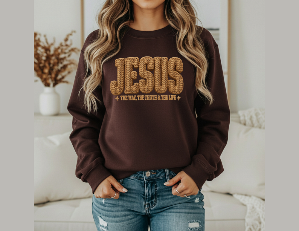 Person wearing Jesus Crew dark brown sweatshirt with mustard-yellow lettering, hands in pockets of light-wash jeans, standing in a cozy living room.