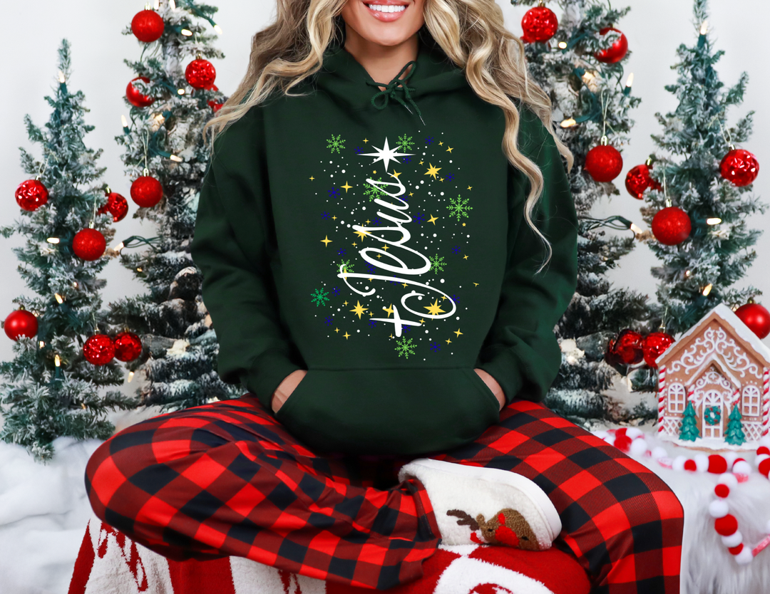 Person in a Jesus Tree Hoodie sits on a festive blanket, surrounded by snow-dusted trees and gingerbread decor, embodying cozy Christmas spirit.