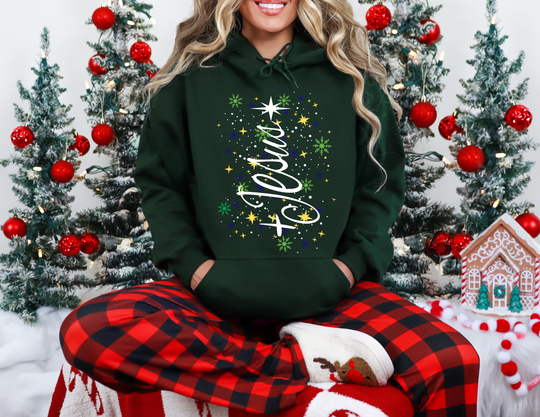 Person in a Jesus Tree Hoodie sits on a festive blanket, surrounded by snow-dusted trees and gingerbread decor, embodying cozy Christmas spirit.