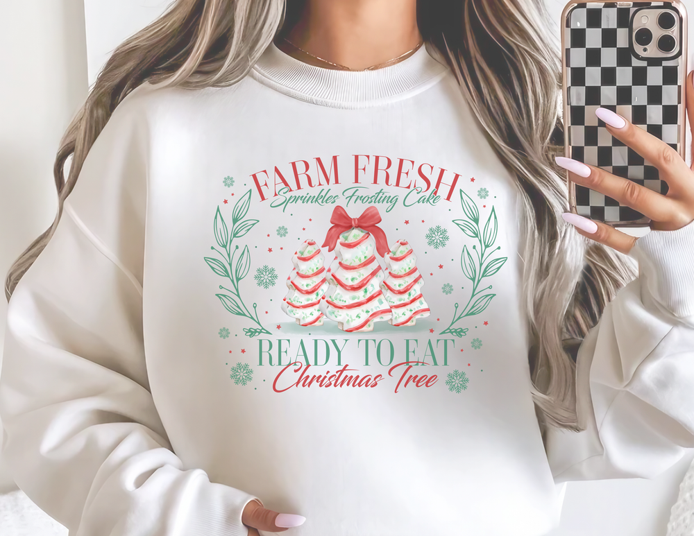 Person in mirror selfie wearing a Farm Fresh Cookies Crew oversized sweatshirt with festive Christmas cake graphics, holding a checkered smartphone case.