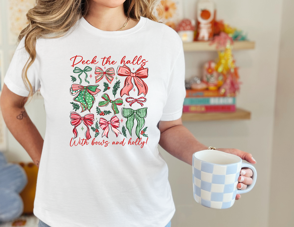 Person wearing 'Deck The Halls Tee' with holiday bows and holly, holding a ceramic mug. Cozy and festive design from Faith Family Designs.